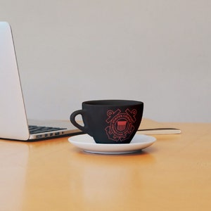 May include: A black coffee mug with a red United States Coast Guard emblem on the side. The mug is sitting on a white saucer on a wooden table.