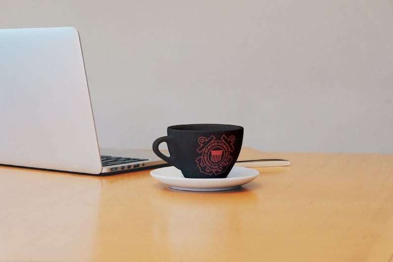 May include: A black coffee mug with a red United States Coast Guard emblem on the side. The mug is sitting on a white saucer on a wooden table.