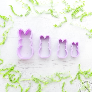 May include: Set of four purple plastic bunny cookie cutters of different sizes. The cutters are on a white wooden surface with green paper shreds around the edges. The cutters are for making bunny-shaped cookies.