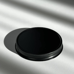 Op de afbeelding: Een ronde, zwarte metalen deksel met een getextureerde rand, rustend op een wit oppervlak. Het oppervlak van de deksel is glanzend en reflecterend. Natuurlijk licht creëert schaduwen.