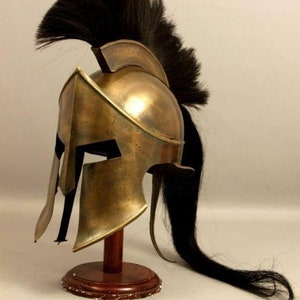 May include: A bronze-colored Spartan helmet with a black plume, displayed on a wooden stand. The helmet features cheek guards and a curved design, showcasing historical craftsmanship. The black plume extends down the back of the helmet.
