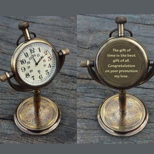 May include: A brass desk clock with a round face and a stand. The clock face has Roman numerals and a minute track. The back of the clock has an inscription that reads: "The gift of time is the best gift of all. Congratulation on your promotion my love."