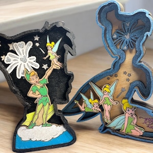 May include: Two wooden figurines of Peter Pan and Tinkerbell. The Peter Pan figurine is black and blue with a cutout of Peter Pan flying with Tinkerbell. The Tinkerbell figurine is brown and blue with a cutout of Tinkerbell flying with a wand. Both figurines have a cutout of fireworks in the background.