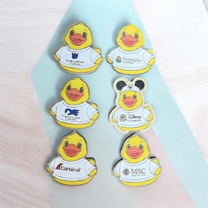 May include: Six yellow rubber duck-shaped wooden ornaments, each wearing a white t-shirt printed with different cruise line logos including Royal Caribbean, Norwegian, Princess, Disney, Carnival, and MSC. The Disney duck features black mouse ears.