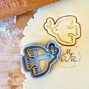 May include: A close-up of a wooden rolling pin and a cookie cutter in the shape of a dove with an olive branch. The cookie cutter is on top of a sheet of cookie dough. The cookie cutter is labelled "My Do Pe" and "Family Made".