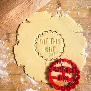 K&ouml;nnte beinhalten: Nahaufnahme eines Holz-Nudelholzes und eines Ausstechers auf einer Holzoberfl&auml;che. Der Ausstecher ist rot und sagt "My Birthday This" und der andere Ausstecher ist auf einem Teigst&uuml;ck und sagt "Eat This Shit Cookie Cutter".