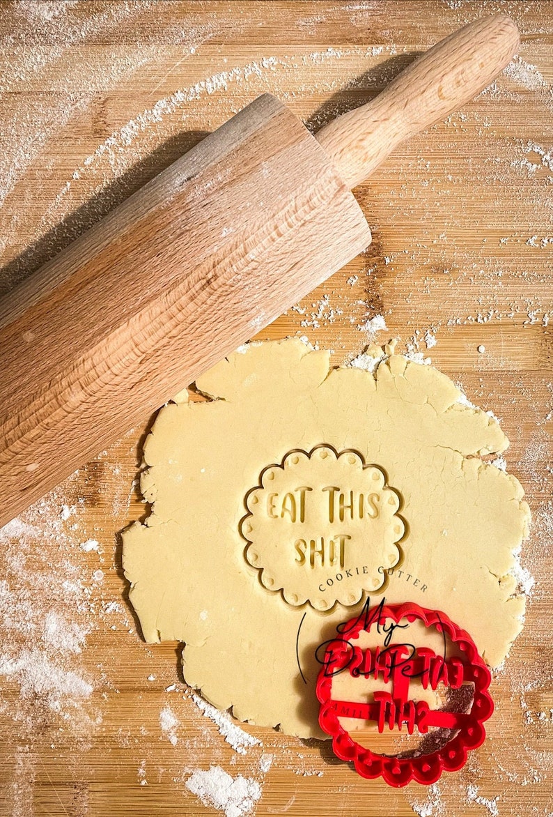 K&ouml;nnte beinhalten: Nahaufnahme eines Holz-Nudelholzes und eines Ausstechers auf einer Holzoberfl&auml;che. Der Ausstecher ist rot und sagt "My Birthday This" und der andere Ausstecher ist auf einem Teigst&uuml;ck und sagt "Eat This Shit Cookie Cutter".