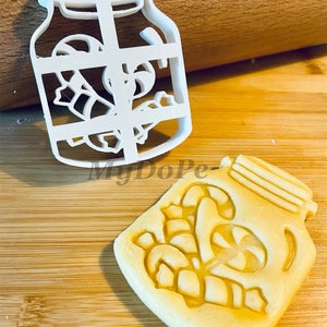 May include: White plastic cookie cutter shaped like a mason jar with candy canes inside. A cookie is also shown that has been cut out using the cookie cutter.