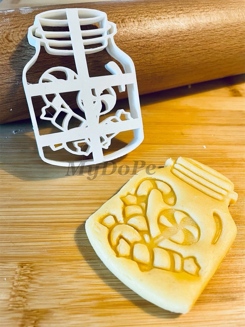 May include: White plastic cookie cutter shaped like a mason jar with candy canes inside. A cookie is also shown that has been cut out using the cookie cutter.