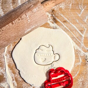 Può includere: Tagliapasta in plastica rossa a forma di tazza con un cuore e il testo "Cookie Monster". Il tagliapasta è su una superficie di legno ricoperta di farina bianca. Sono visibili anche un mattarello e un cerchio di pasta.