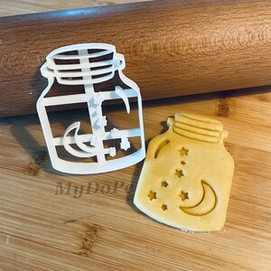 May include: White plastic cookie cutter shaped like a mason jar with a crescent moon and stars inside. A cookie cut with the same design is shown next to the cutter.