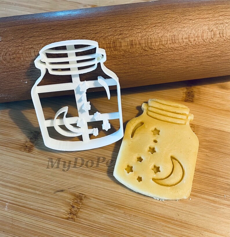 May include: White plastic cookie cutter shaped like a mason jar with a crescent moon and stars inside. A cookie cut with the same design is shown next to the cutter.