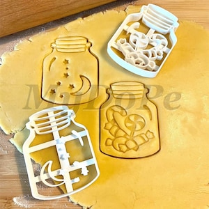 May include: Three white plastic cookie cutters shaped like mason jars with different designs. The cutters are on a sheet of yellow cookie dough.
