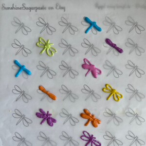 May include: A sheet of parchment paper with outlined dragonfly shapes.  The outlines are filled in with various colors of sugarpaste, including blue, green, yellow, orange, pink, and purple.