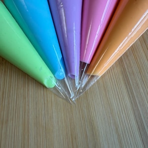 May include: Five clear plastic piping bags filled with icing in vibrant colors arranged in a fan shape. The colors include light green, bright blue, lavender purple, soft pink, and light orange. Each bag is filled to the top and sealed.