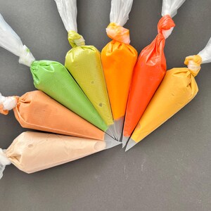 May include: Six piping bags filled with different colored frosting, arranged in a fan shape on a gray surface. The colors are peach, green, yellow, orange, red, and yellow.