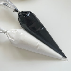 May include: Two piping bags, one black and one white, are lying on a white surface. The bags are filled with frosting or other decorating material.