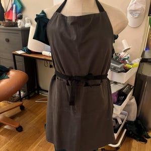 May include: A dark grey apron with black straps and a waist tie, shown on a mannequin. The apron features two front pockets and a straightforward design. The apron is made of a robust fabric, suitable for kitchen or workshop use.