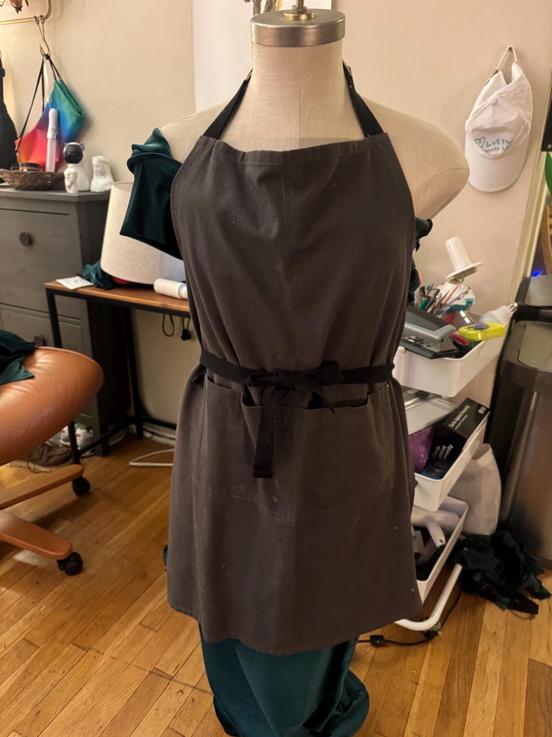 May include: A dark grey apron with black straps and a waist tie, shown on a mannequin. The apron features two front pockets and a straightforward design. The apron is made of a robust fabric, suitable for kitchen or workshop use.