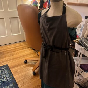 May include: A brown apron with a black strap and waist tie is displayed on a mannequin. The apron is made of a dark fabric and has a simple design. The mannequin is wearing a teal skirt. The background includes a wooden floor and a chair.