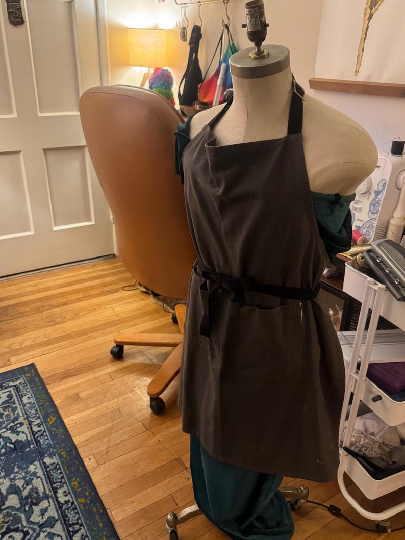 May include: A brown apron with a black strap and waist tie is displayed on a mannequin. The apron is made of a dark fabric and has a simple design. The mannequin is wearing a teal skirt. The background includes a wooden floor and a chair.