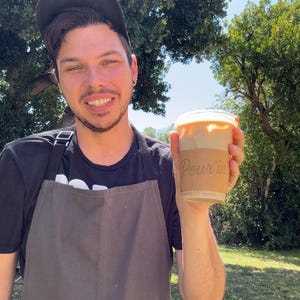 May include: A person holding a clear plastic cup of iced coffee with a cardboard sleeve that reads "Pour'in". The person is wearing a black apron and a black baseball cap. The background is a sunny outdoor scene with trees.