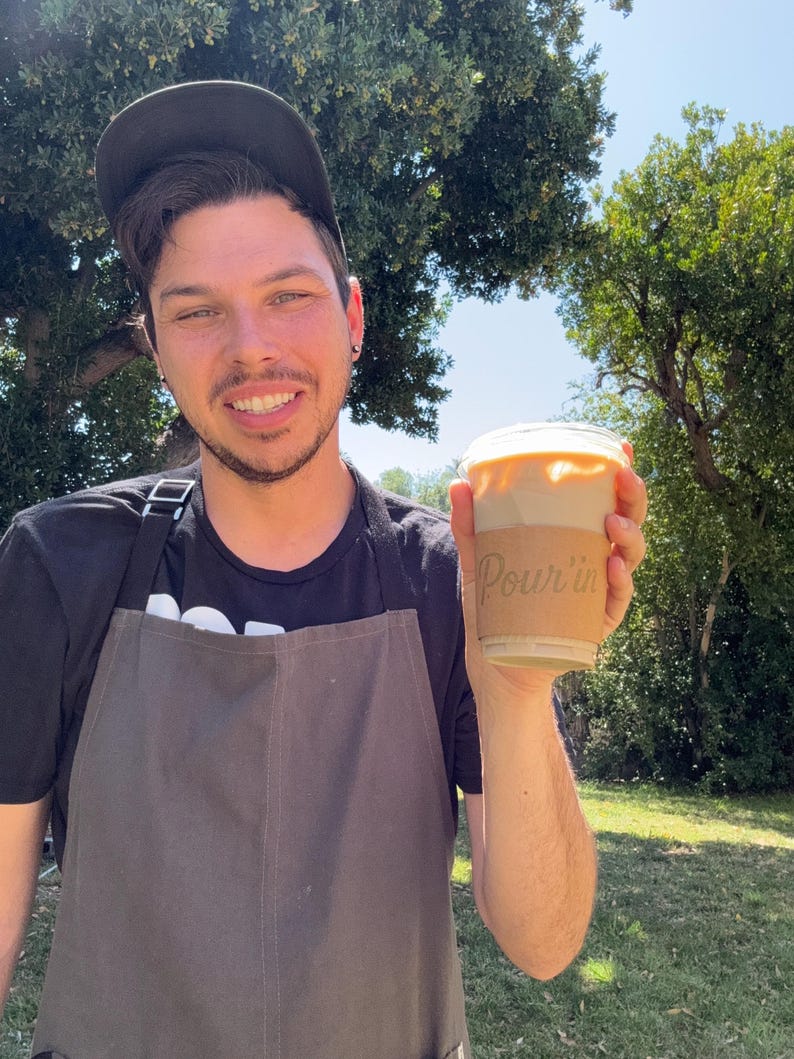 May include: A person holding a clear plastic cup of iced coffee with a cardboard sleeve that reads "Pour'in". The person is wearing a black apron and a black baseball cap. The background is a sunny outdoor scene with trees.