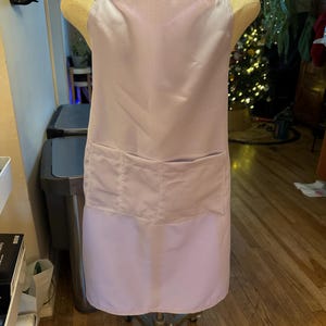 May include: A light purple apron with a large front pocket and black straps. The apron is displayed on a mannequin. The apron is made of a smooth, satin-like material.