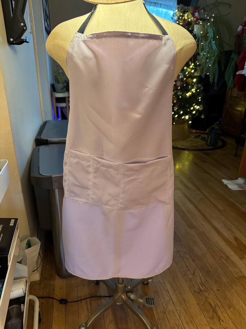 May include: A light purple apron with a large front pocket and black straps. The apron is displayed on a mannequin. The apron is made of a smooth, satin-like material.