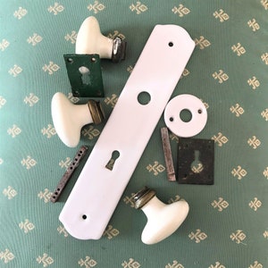 May include: A collection of vintage door hardware components, including white porcelain doorknobs with brass accents, a white door plate, and various metal pieces. The hardware is arranged on a patterned green fabric.