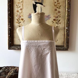 Può includere: Un abito bianco senza maniche con scollo quadrato e spalline sottili. La parte superiore del petto è decorata con intricati ricami floreali bianchi e motivi circolari traforati, esposto su un manichino in tessuto color beige.
