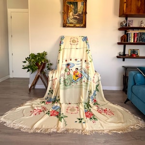 Op de afbeelding: Een crèmekleurige, rechthoekige plaid met een centrale illustratie van twee figuren in traditionele kleding, de een met een paraplu en de ander met een snaarinstrument, omringd door bloemmotieven, rode geometrische randen en franjes.