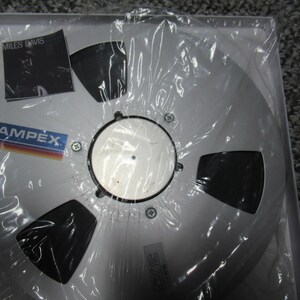 K&ouml;nnte beinhalten: Ein silbernes Tonband mit einem schwarzen Etikett. Das Etikett tr&auml;gt den Text "MILES DAVIS" und das Logo "AMPEX".
