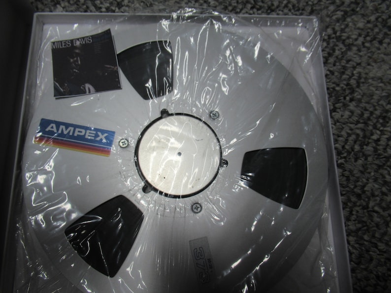 K&ouml;nnte beinhalten: Ein silbernes Tonband mit einem schwarzen Etikett. Das Etikett tr&auml;gt den Text "MILES DAVIS" und das Logo "AMPEX".