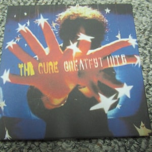 May include: The Cure Greatest Hits CD cover featuring a red hand with a blue and white starry background. The text "The Cure Greatest Hits" is visible in yellow.