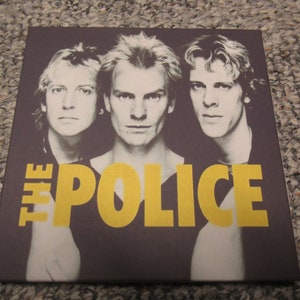 May include: A black and white photo of The Police band members, Sting, Andy Summers, and Stewart Copeland. The band's name is written in yellow letters across the bottom of the photo.