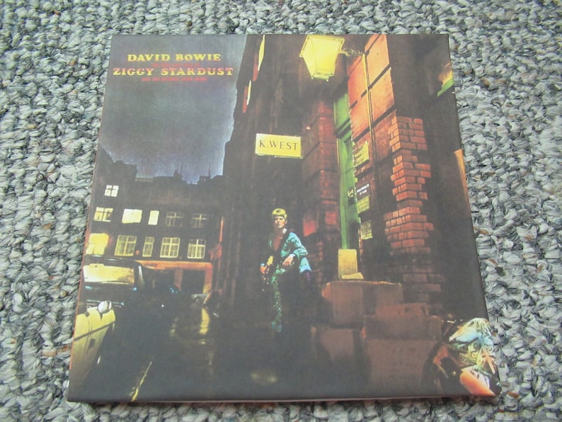 May include: Album cover for David Bowie's "Ziggy Stardust" depicting a nighttime street scene with a figure in a colourful outfit. The album title and artist name are visible at the top. The scene includes brick buildings, streetlights, and the text "K. West".