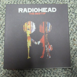 Puede incluir: Una caja de CD negra con el texto "Radiohead The Best Of" en blanco. La portada presenta una silueta de dos personas hechas de patrones abstractos coloridos.