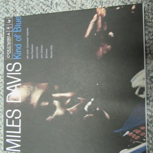 K&ouml;nnte beinhalten: Ein schwarzes und wei&szlig;es Vinyl-Plattencover mit dem Titel "Kind of Blue" von Miles Davis. Das Cover zeigt eine Nahaufnahme von Miles Davis' Gesicht und Hand, die eine Trompete h&auml;lt.