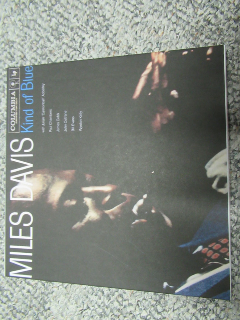 K&ouml;nnte beinhalten: Ein schwarzes und wei&szlig;es Vinyl-Plattencover mit dem Titel "Kind of Blue" von Miles Davis. Das Cover zeigt eine Nahaufnahme von Miles Davis' Gesicht und Hand, die eine Trompete h&auml;lt.