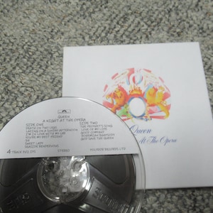 Puede incluir: Una cinta de carrete Queen "A Night at the Opera" y la portada del álbum. La portada del álbum es blanca con el escudo de la banda y el título del álbum. La cinta tiene listas de canciones y el logotipo de Polygram Records.