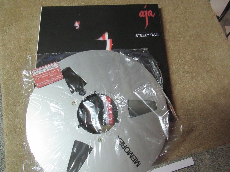 Puede incluir: Una bobina de cinta de audio Memorex plateada en una funda de pl&aacute;stico, con una portada de &aacute;lbum negra detr&aacute;s. La portada del &aacute;lbum es negra con la palabra "aja" en escritura roja y "Steely Dan" en texto blanco.