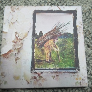 May include: A vintage album cover with a faded white background and a framed image of a man carrying a bundle of sticks on his back. The man is wearing a brown jacket and pants and is walking through a field.