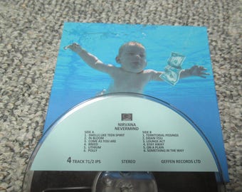 Puede incluir: Una cinta de casete con una portada azul y blanca que muestra una foto de un niño pequeño nadando bajo el agua. La cinta está etiquetada como "Nirvana Nevermind" y enumera las canciones de cada lado.