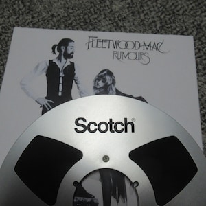 Puede incluir: Una cinta de carrete a carrete plateada con la palabra "Scotch" impresa en ella. La cinta est&aacute; encima de una portada de &aacute;lbum blanca con el texto "Fleetwood Mac Rumours".