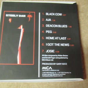 Puede incluir: Caja de CD del &aacute;lbum "Aja" de Steely Dan. La portada es negra con un dise&ntilde;o abstracto en rojo y blanco. La contraportada muestra la lista de canciones, con t&iacute;tulos y duraciones en rojo.