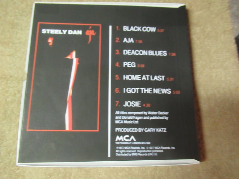 Puede incluir: Caja de CD del &aacute;lbum "Aja" de Steely Dan. La portada es negra con un dise&ntilde;o abstracto en rojo y blanco. La contraportada muestra la lista de canciones, con t&iacute;tulos y duraciones en rojo.