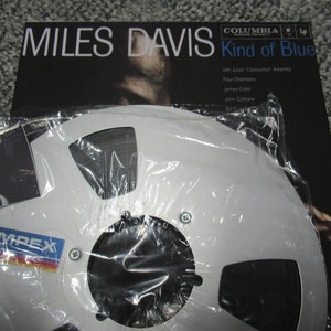 K&ouml;nnte beinhalten: Ein wei&szlig;es Tonband mit schwarzem Etikett und dem Text "AMPEX" in Blau. Das Band befindet sich in einer durchsichtigen Plastikt&uuml;te und liegt neben einem Vinyl-Plattencover f&uuml;r Miles Davis' "Kind of Blue".