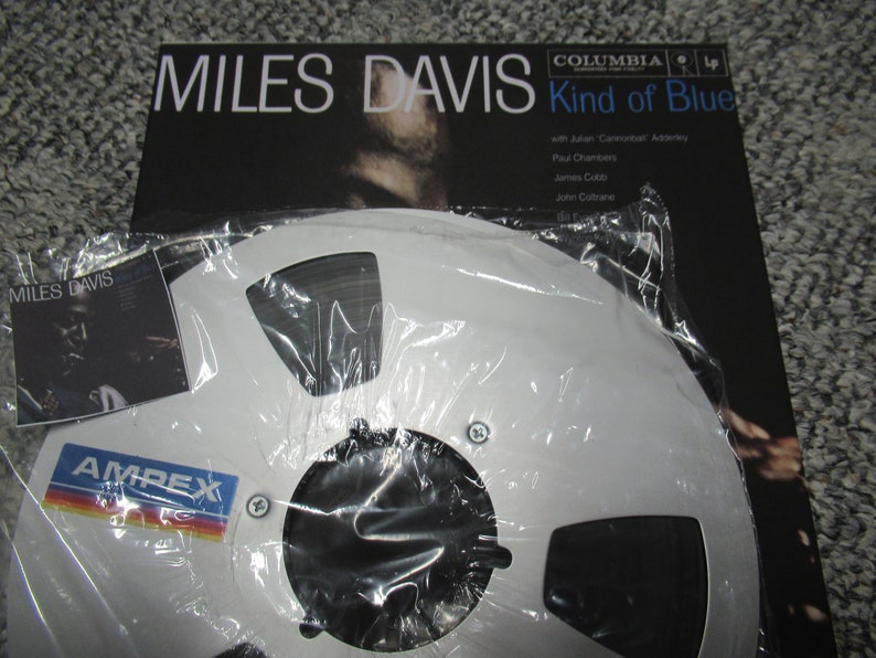 K&ouml;nnte beinhalten: Ein wei&szlig;es Tonband mit schwarzem Etikett und dem Text "AMPEX" in Blau. Das Band befindet sich in einer durchsichtigen Plastikt&uuml;te und liegt neben einem Vinyl-Plattencover f&uuml;r Miles Davis' "Kind of Blue".