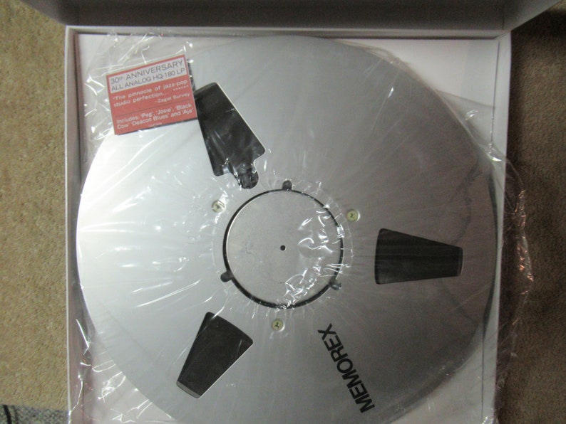 Puede incluir: Una cinta de carrete a carrete Memorex plateada en una caja blanca, envuelta en pl&aacute;stico transparente. La cinta tiene un cubo central con recortes y est&aacute; etiquetada como "30th Anniversary All Analog HQ 180 LP". Una peque&ntilde;a etiqueta enumera las canciones incluidas.